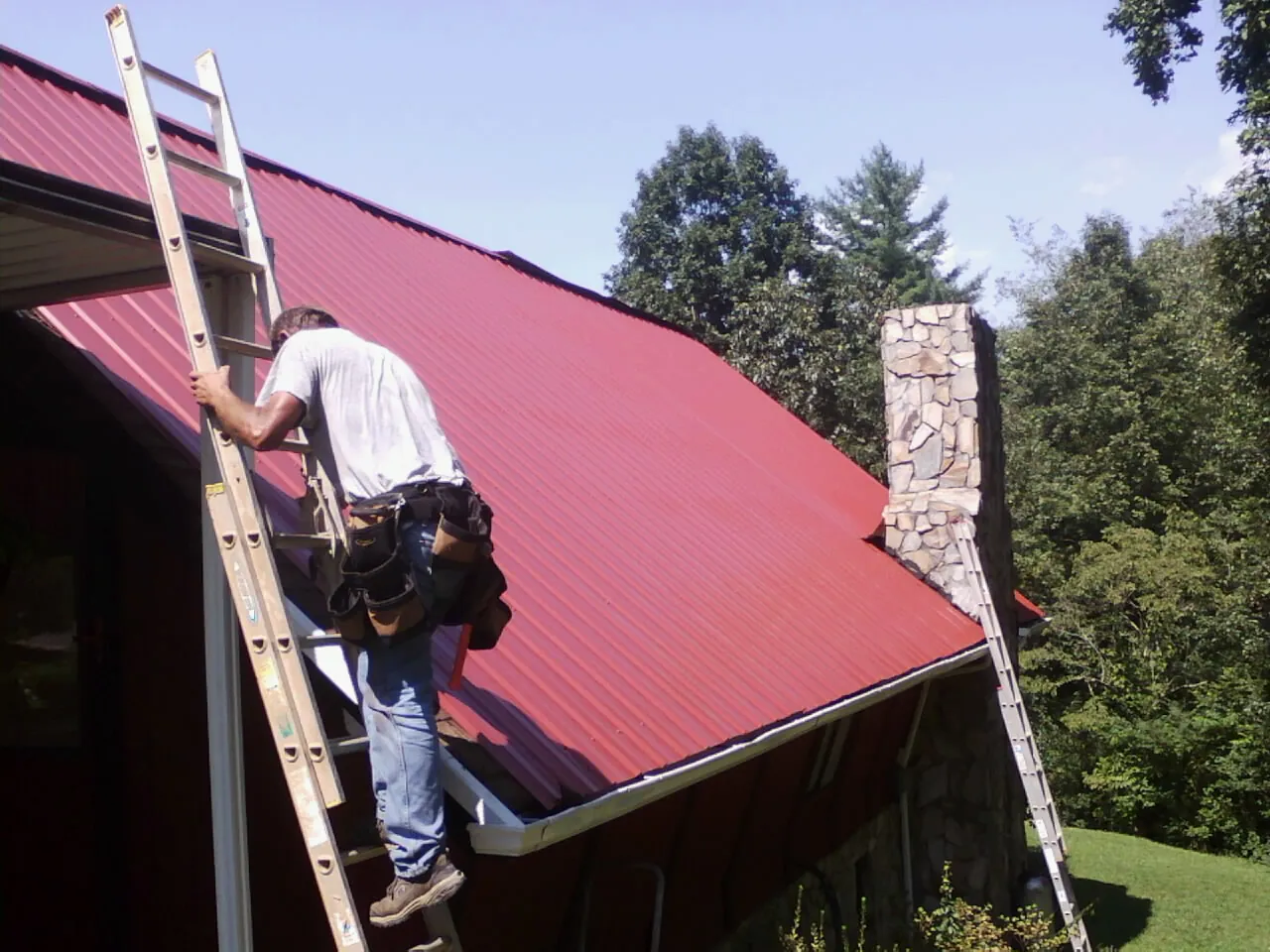 Roofer on a ladder beside a red metal roof and stone chimney — Soffit & Fascia Repair by Roofing Brothers in Howells